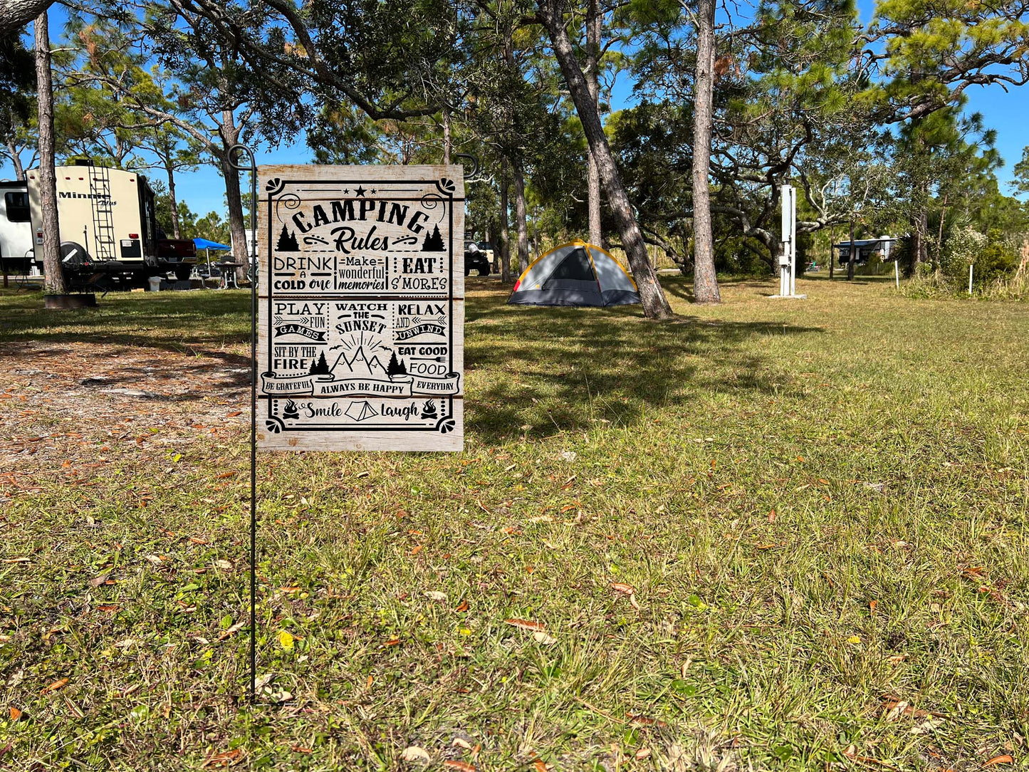 'Camping Rules' garden flag on garden flag stand in a grassy area with trees and campers at a campground in the background
