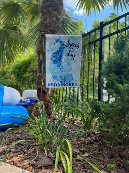 Personalized garden flag text read ' Seas the Day' with a seahorse design in a garden setting in front of a palm tree and pool toys