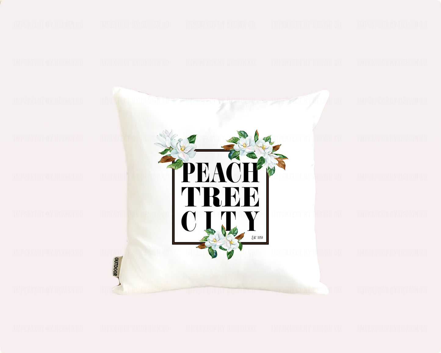 16x16 inches An elegant white outdoor pillow with a weather-resistant poly coating, featuring a stunning magnolia bloom design and the words "Peachtree City est. 1959" inside a black frame | imperfect by design co imperfect by design co