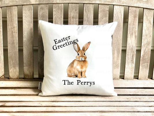 White personalized indoor outdoor pillow advertising pillow cover with Easter bunny design and text reading 'Easter Greetings - The Perrys' text on a wooden porch bench.