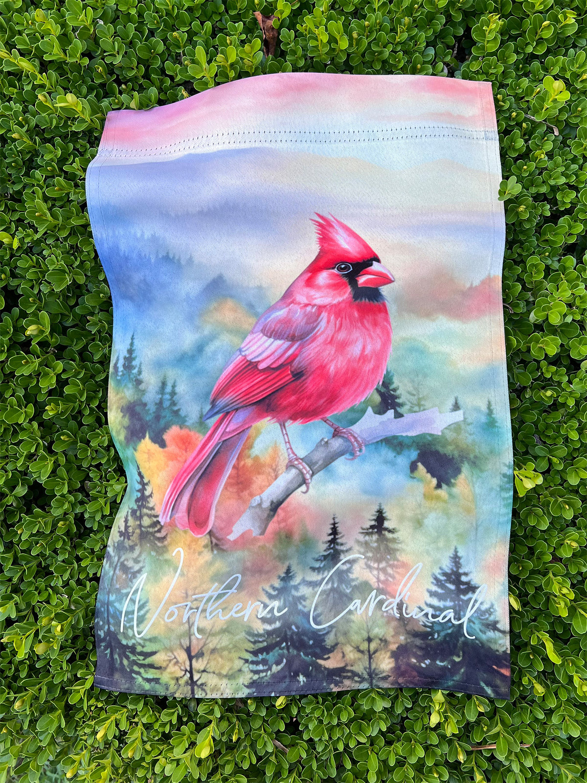 12x18 Garden Flag with a large red cardinal perched on a branch against a painted forest background, placed on a boxwood hedge. The garden flag has a pastel pink and blue top border and the words 'Northern Cardinal' printed near the bottom. | imperfect by design co