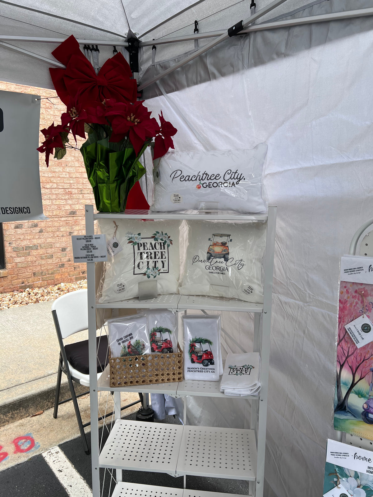 A white metal display shelf under a white tent showcasing 'Peachtree City Souvenirs including printed pillowcases, small packaged items, and a large red poinsettia arrangement on top.
