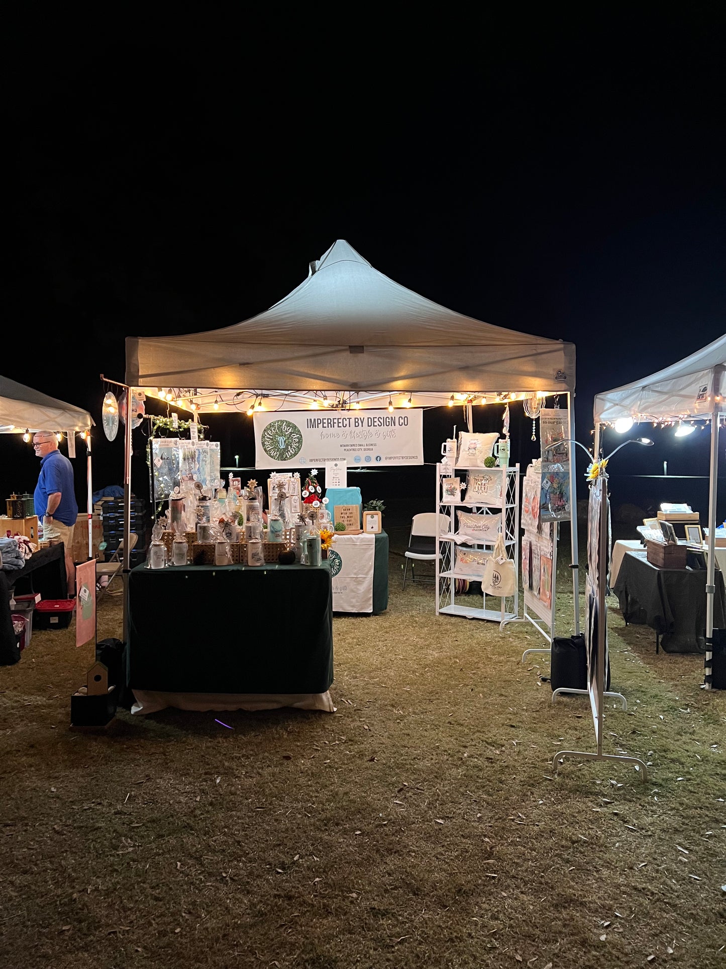 Outdoor Vendor Market setting with a tent and tables under a dark sky. Tent is illuminated with string lights. Banner in back of tent reads 'imperfect by design co' and features the imperfect by design co logo Tables with green table cloths feature a variety of imperfect by design co products drinkware etc. Garden flags and pillow are visible are the background