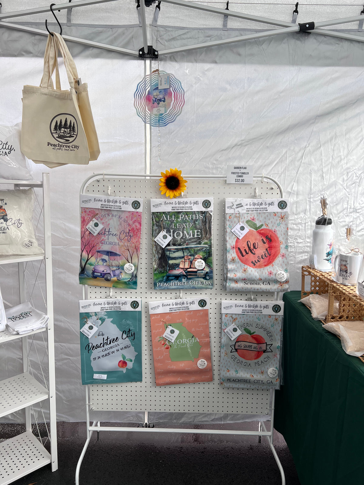 Collection of Peachtree City garden flags and tote bags on a table with a garden flag display in a vendor tent. imperfect by design co