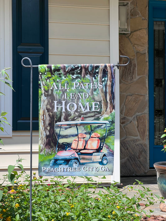 Garden flag with golf cart and 'All Paths Lead Home' text in front of a house on a garden flag stand. The flag depicts a brown golf cart parked on a windy tree lined path. Text placed at bottom of flag reads Peachtree City. imperfect by design co exclusive design