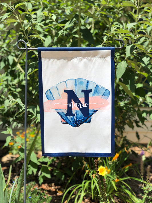 A personalized garden flag with a navy blue border, a blue seashell graphic, and the name 'Nario' in white script, displayed on a metal stand in a garden setting.
