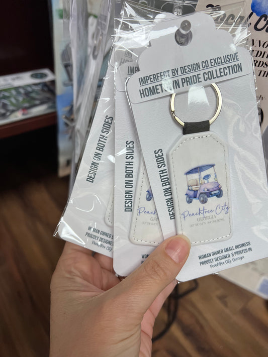 Several retail packaged white faux leather keychain with a blue golf cart and purple text, including coordinates, and the text 'Peachtree City, GA'. On a retail stand held up by a hand | imperfect by design co exclusive design