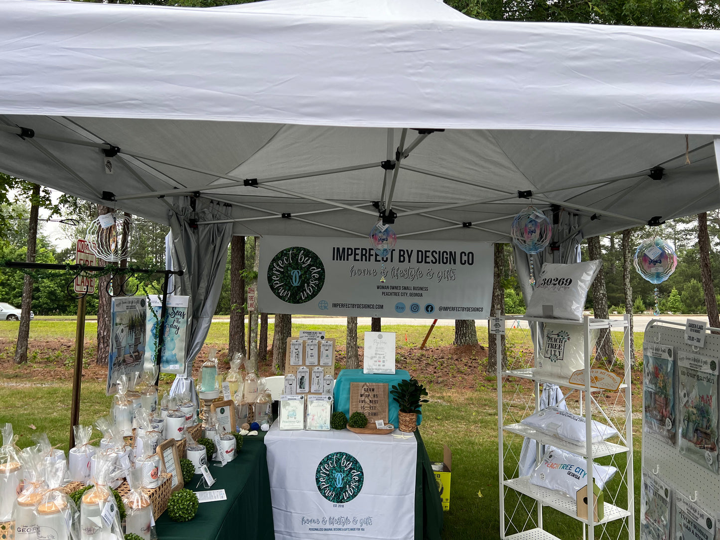 Product display at Peachtree City Vendor Market featuring imperfect by design co Products (Wind Spinners, Garden Flags, Coffee Cups, Frosted Tumblers, Key Chains, Notebooks, Pillows Indoor Outdoor