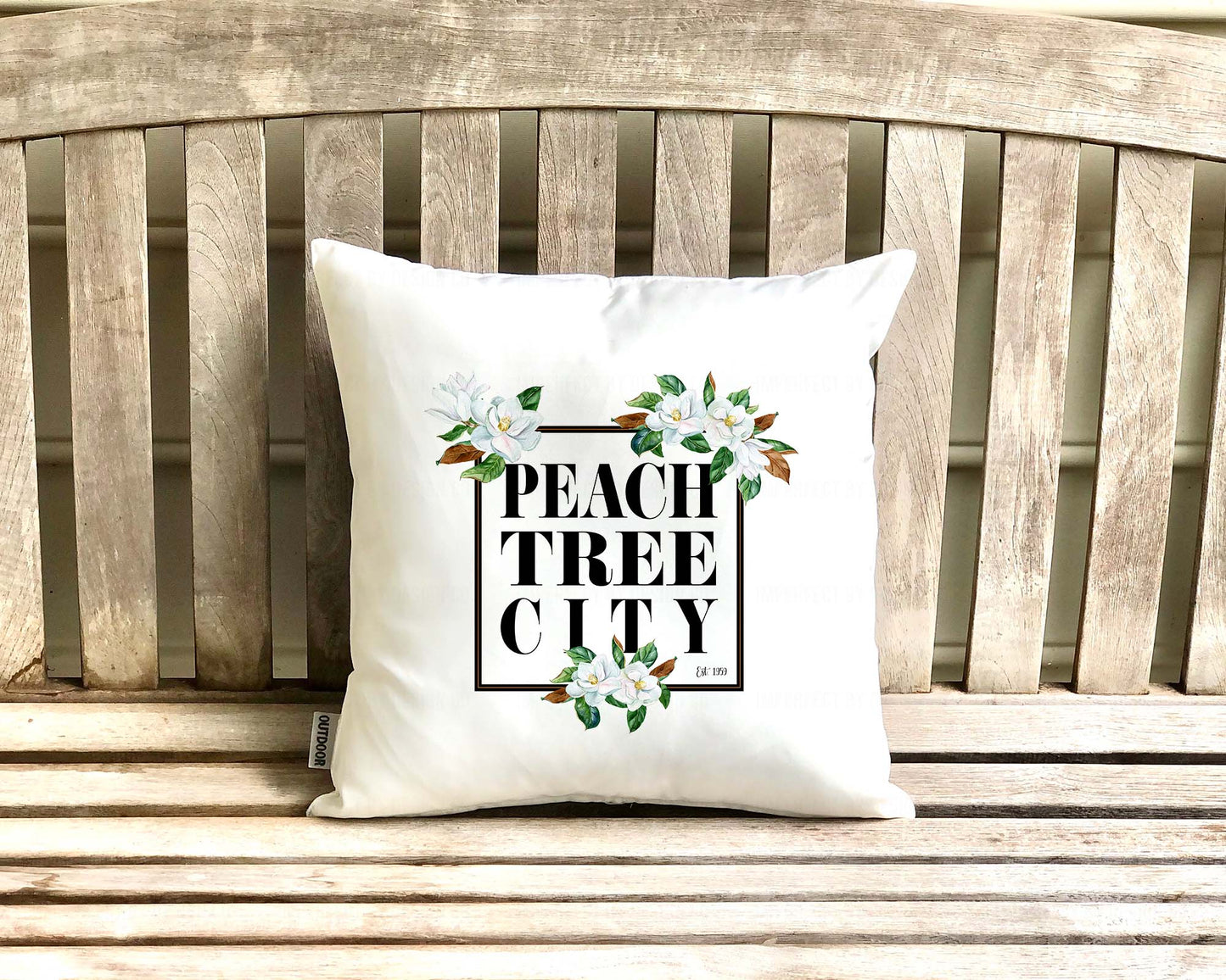 16x16 inches A white outdoor pillow with a black frame adorned with magnolia blooms and the words "Peachtree City est. 1959" inside the frame, perfect for adding comfort and elegance to your outdoor seating area | imperfect by design co