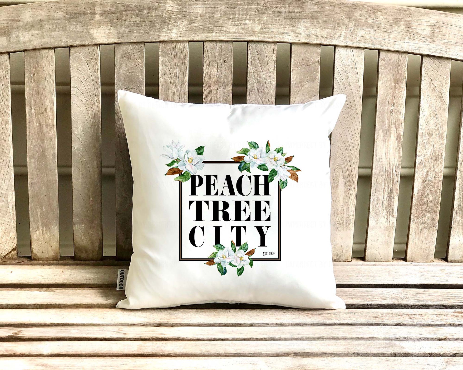 16x16 inches A white outdoor pillow with a black frame adorned with magnolia blooms and the words "Peachtree City est. 1959" inside the frame, perfect for adding comfort and elegance to your outdoor seating area | imperfect by design co
