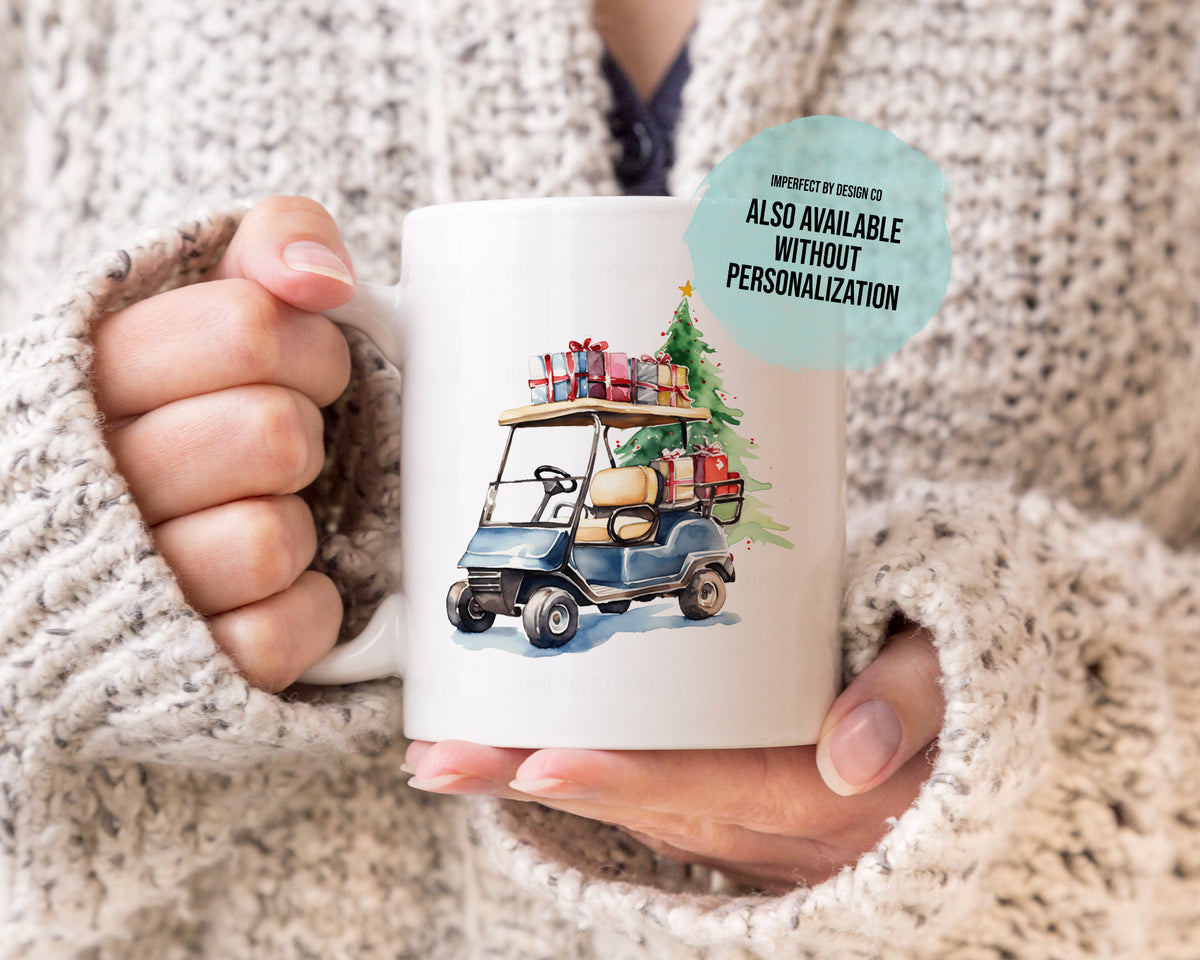 A person in a chunky knit beige sweater holds a white ceramic coffee mug featuring a watercolor illustration of a blue golf cart carrying a Christmas tree and gift boxes.
