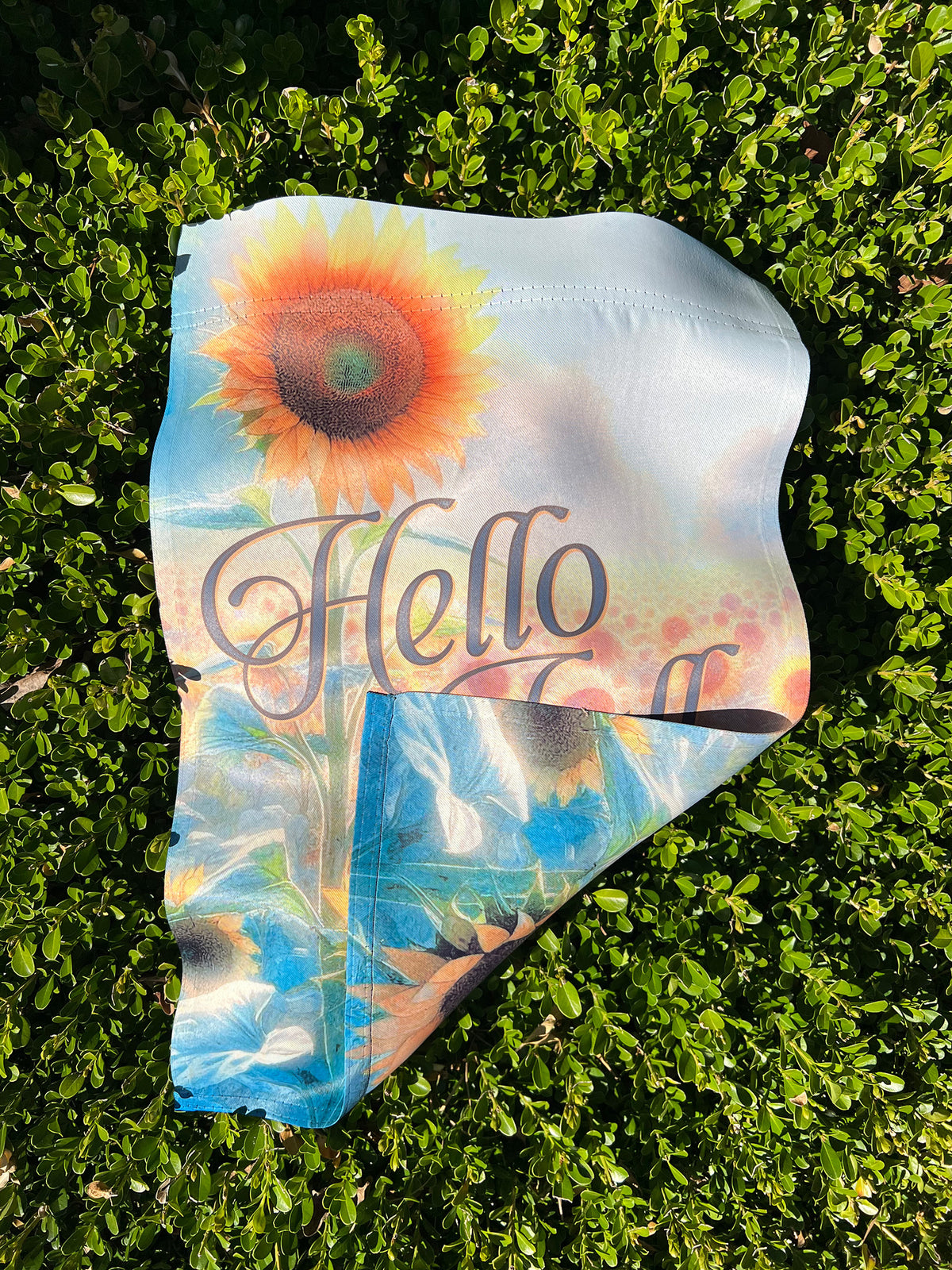 A folded corner on a laid out garden flag  featuring a sunflower and the word 'Hello' in script, placed on a boxwood hedge.  | imperfect by design co
