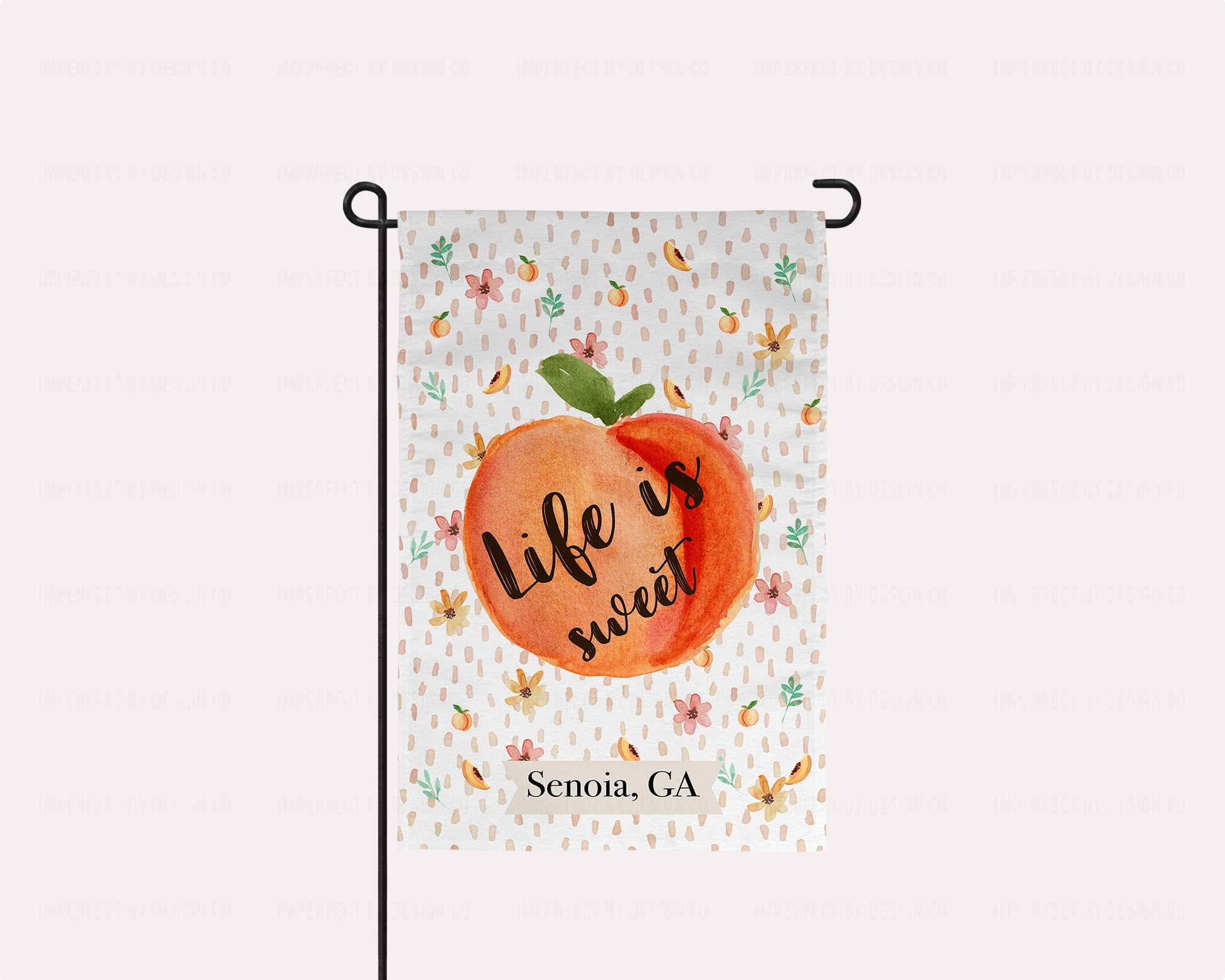 Exclusive imperfect by design co design Personalized decorative garden flag with peach design and text on a white background filled with peaches, peach blooms and leaves. Garden flag is hug on a garden flag pole on a pale pink background. Text on Flag reads 'Life is sweet' and 'Senoia, GA' in black text.