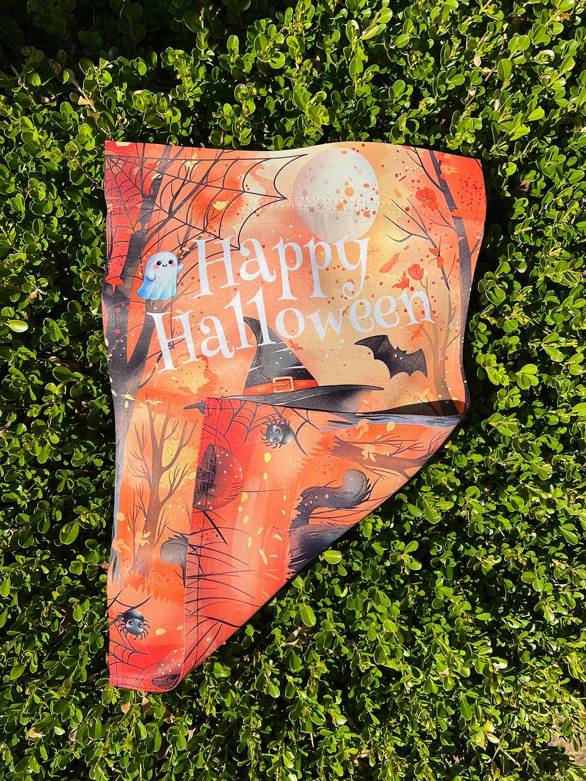 A printed Halloween-themed garden flag laid on boxwood hedge, featuring the words “Happy Halloween,” a full moon, bats, a witch’s hat, and an owl.