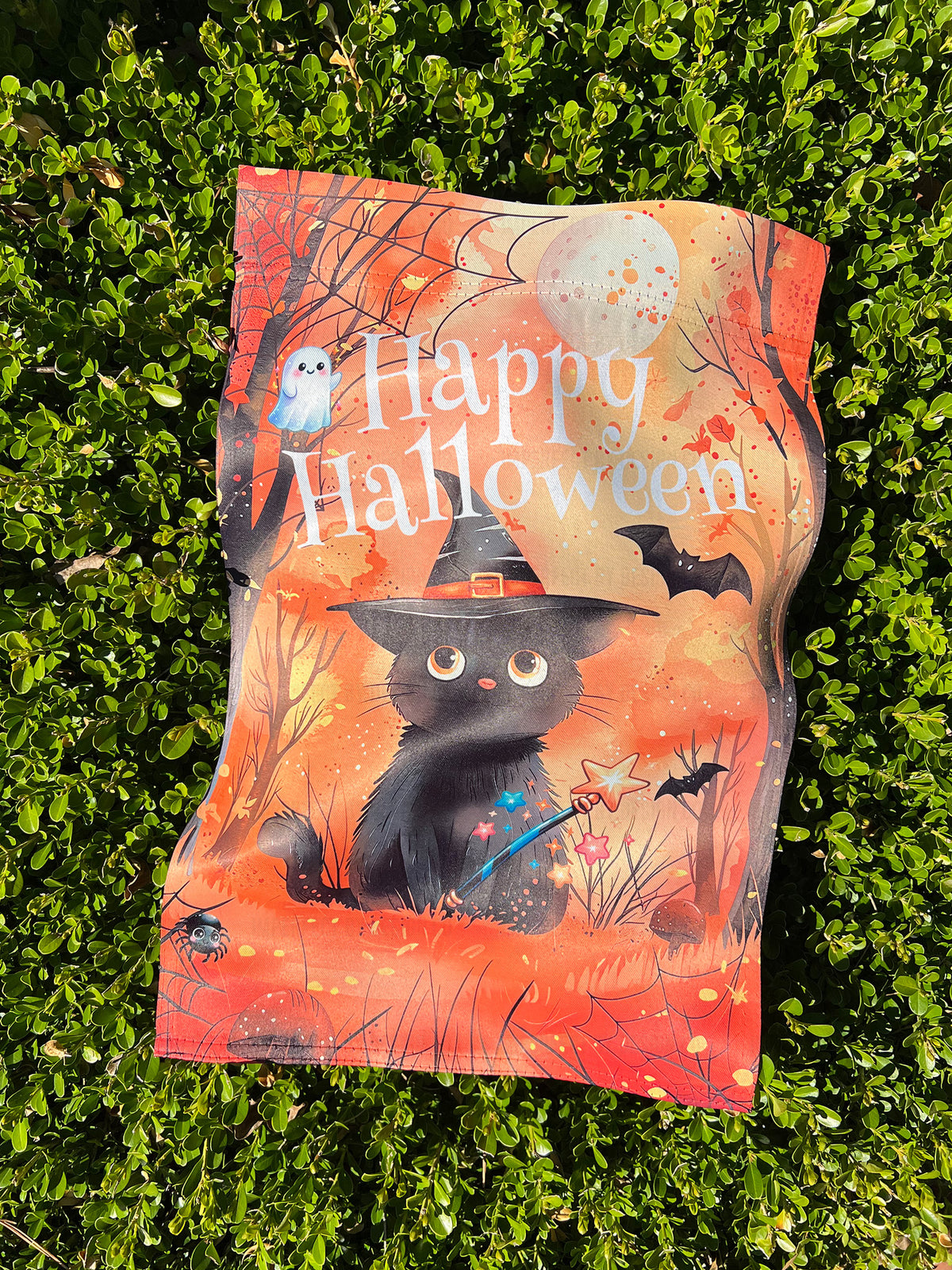 imperfect by design co exclusive 
A Halloween-themed decorative flag with a black cat in a witch hat, bats, a ghost, and the words 'Happy Halloween,' photographed lying on green hedge foliage outdoors.