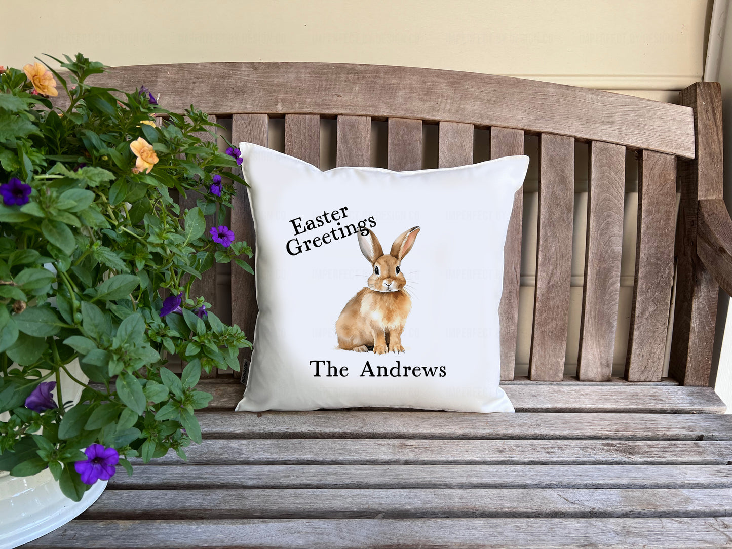 White decorative pillow advertising pillow cover prominently featuring an Easter Bunny with text that reads 'Easter Greetings' on a wooden bench with potted flowers.