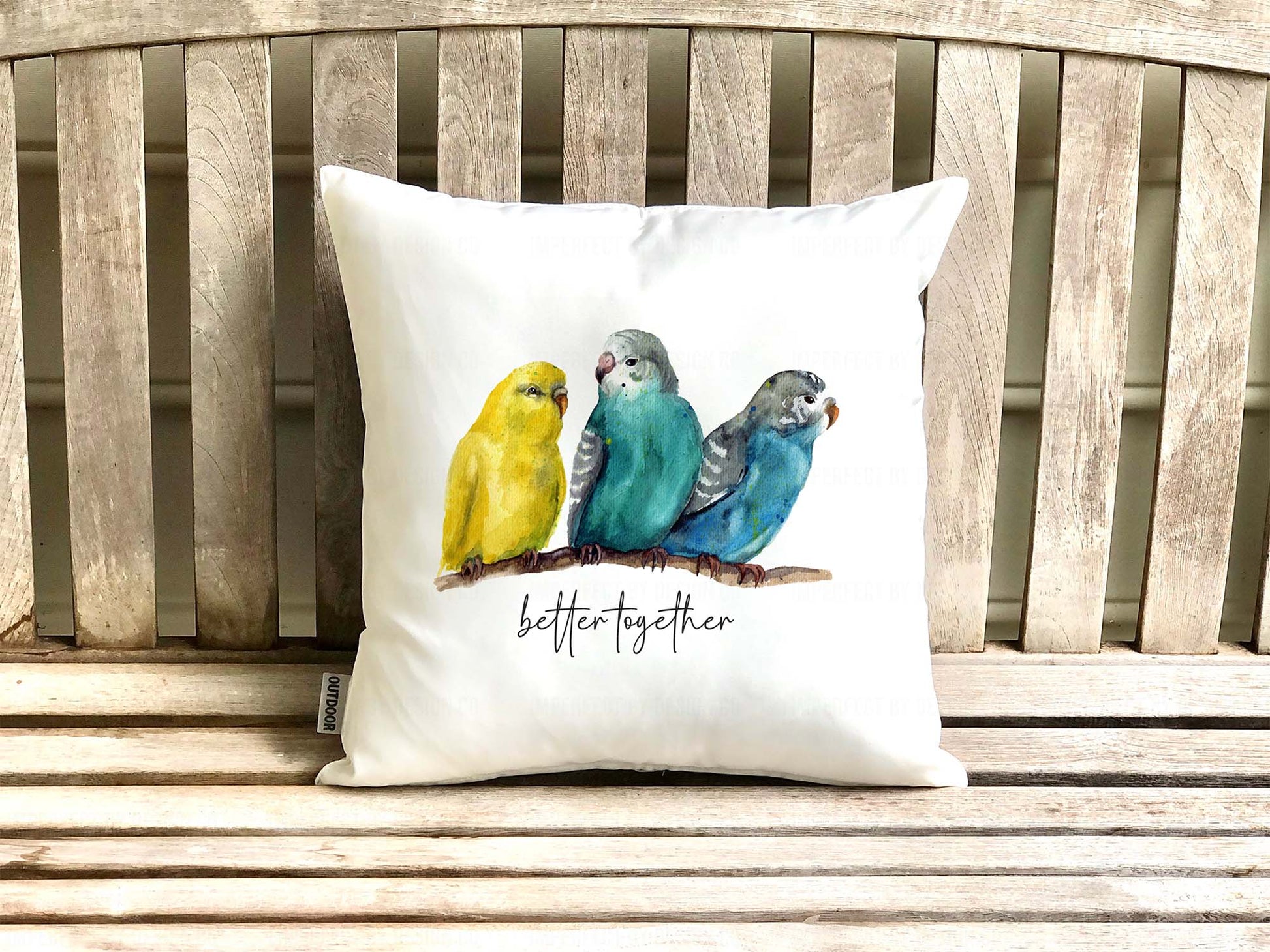 White decorative pillow advertising pillow cover with two watercolor blue budgies and one yellow on sitting on a branch Text on pillow reads 'better together' placed on a wooden bench.