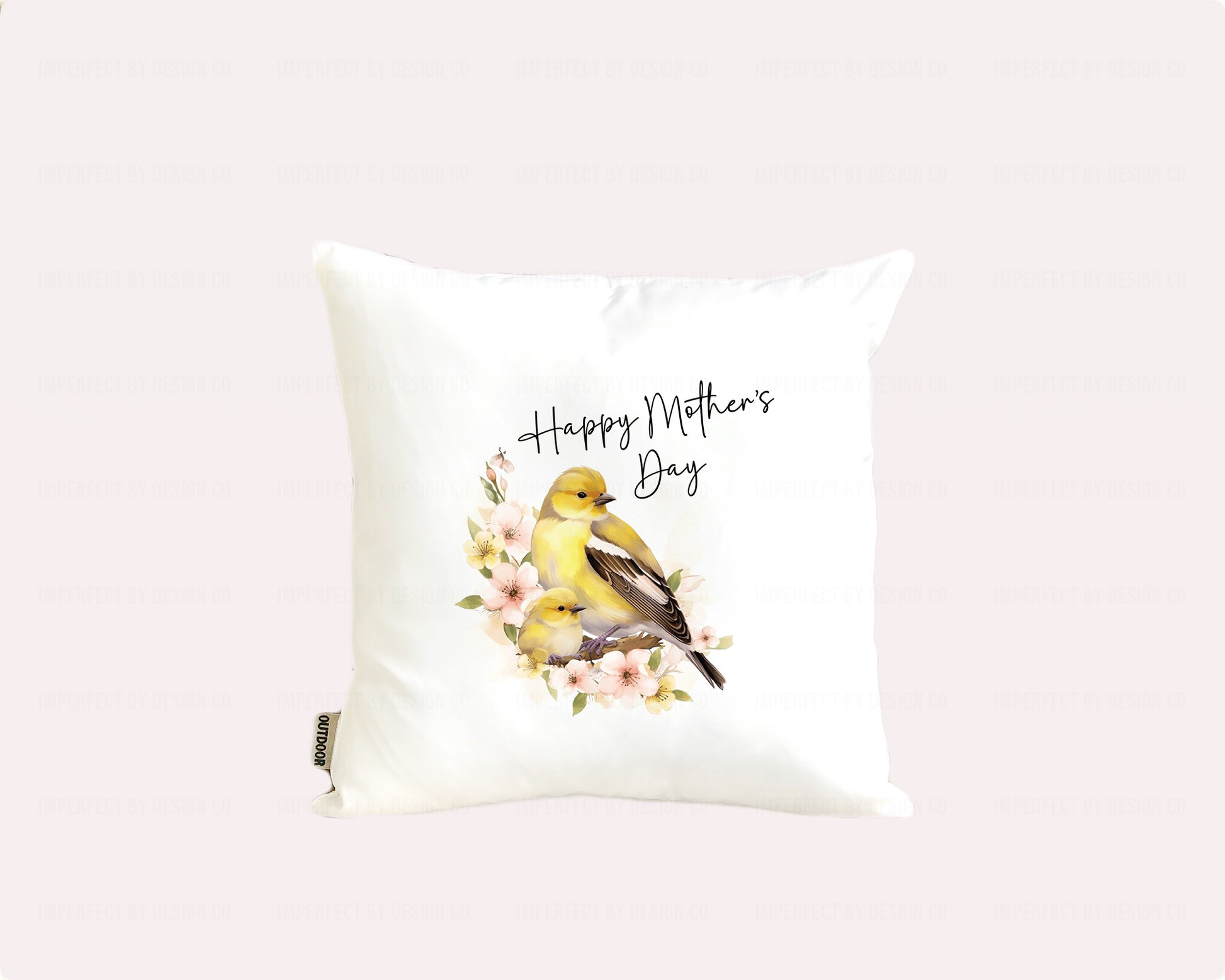 White decorative pillow advertising pillow cover with a female Goldfinch and it's nestling sitting on a branch with pink flowers. Text on pillow reads "Happy Mother's Day' . Placed on a pale pink background  | imperfect by design co