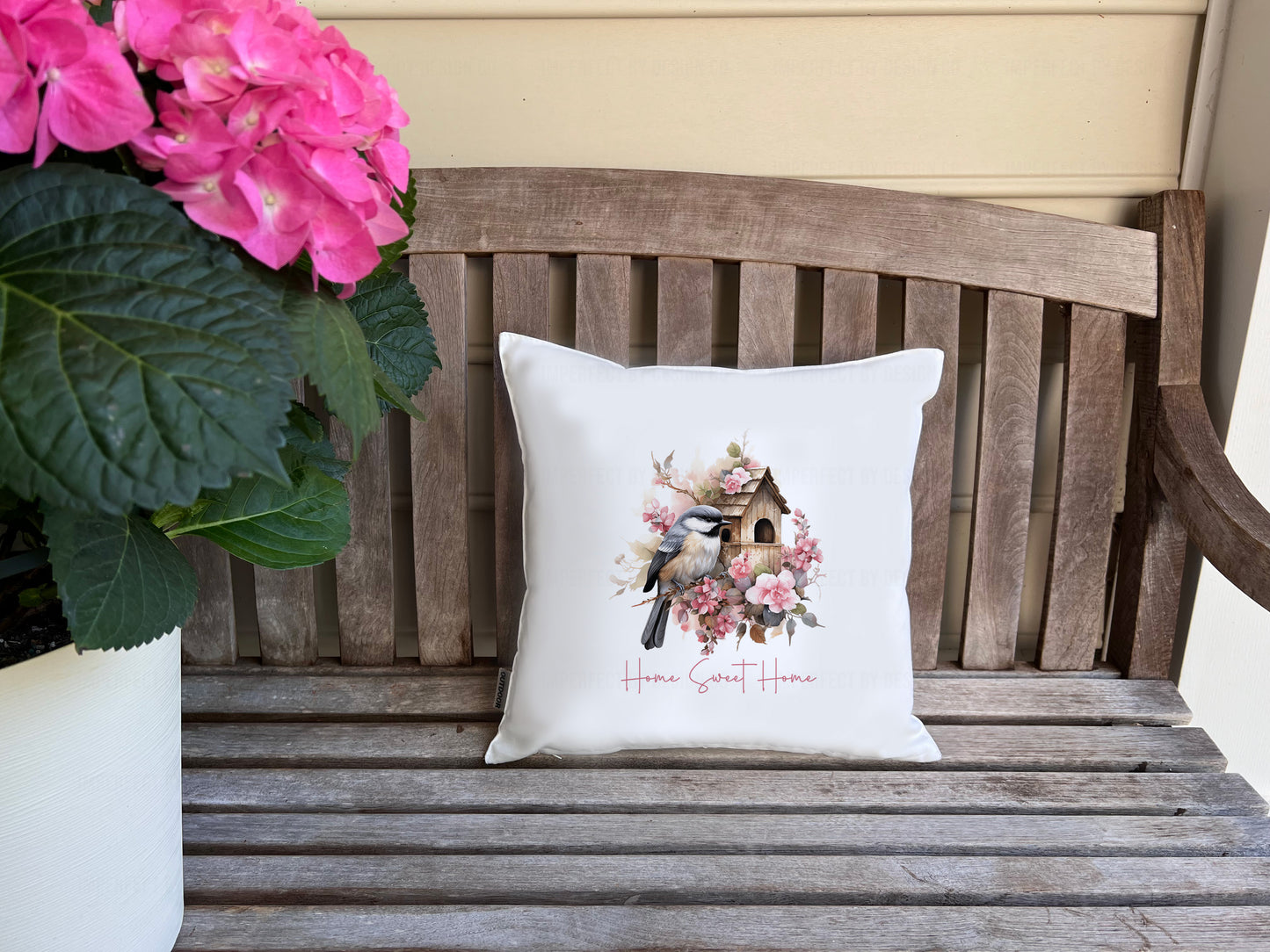 Decorative pillow with a bird and floral design on a wooden bench with pink flowers. Text on pillow cover reads 'Home Sweet Home'  | imperfect by design co