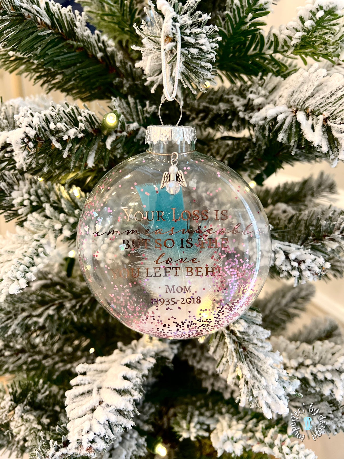 Clear glass ornament with teal-tipped feather, angel charm, and glitter hanging on a tree.