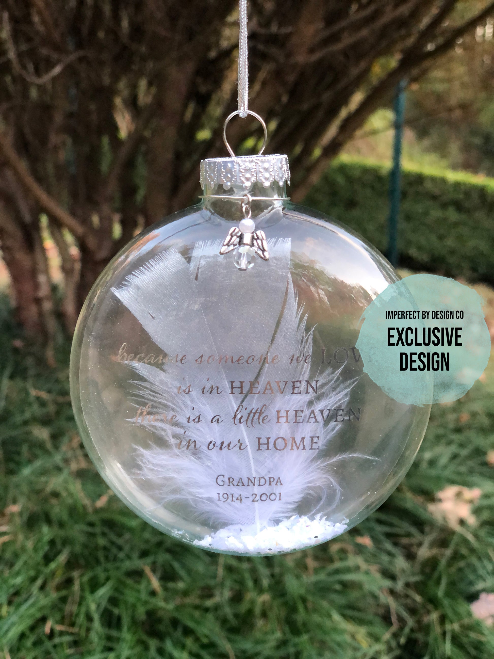 Personalized Memorial Ornament in Clear Glass – A 4.5 x 4 inch disk-shaped clear glass ornament with the text "Because someone we love is in heaven, there is a little heaven in our home," personalized with the person's name and years of life in silver metallic print, featuring a silver-tipped white feather, faux snow, and a small stylized angel charm.