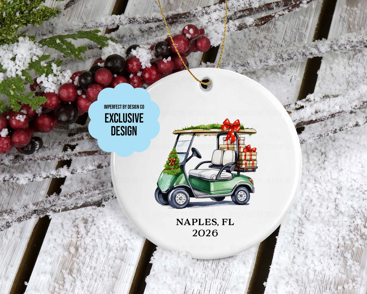 imperfect by design co exclusive design 
A round white ceramic Christmas ornament featuring a green golf cart carrying holiday gifts, with 'NAPLES, FL 2026' printed below. The ornament hangs from a gold string and is styled on a snowy wooden surface with red berries and greenery.