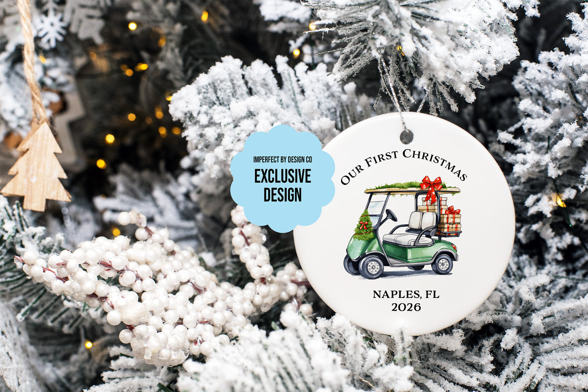imperfect by design co exclusive 
Round white Christmas ornament hanging on a flocked Christmas tree. The ornament features an illustration of a green golf cart carrying wrapped gifts, with the text 'OUR FIRST CHRISTMAS' and 'NAPLES, FL 2026'.