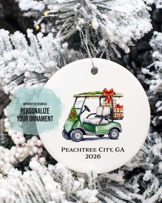 imperfect by design co exclusive design
A round white personalized Christmas ornament hanging on a frosted Christmas tree. The ornament features an illustration of a green golf cart carrying gifts and the text 'PEACHTREE CITY, GA 2026'.
