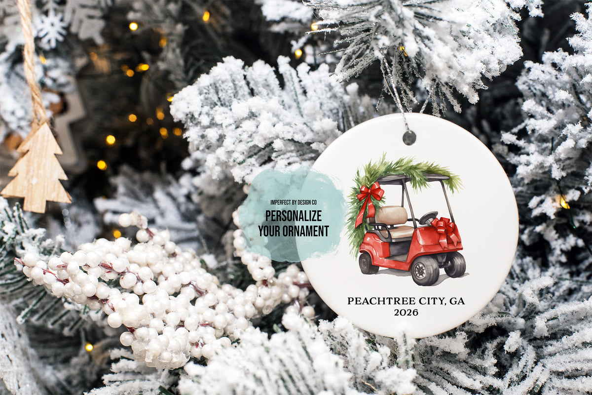 imperfect by design co design exclusive
A round white Christmas ornament featuring an illustration of a red golf cart with green pine garland and a red bow, hanging on a frosted Christmas tree. The ornament includes customizable text reading 'PEACHTREE CITY, GA 2026' and a teal tag that says 'PERSONALIZE YOUR ORNAMENT'.