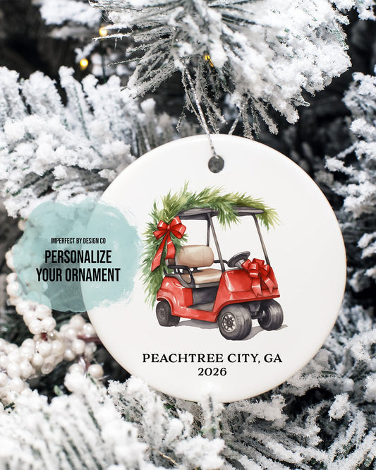 imperfect by design co design exclusive
A round white Christmas ornament featuring an illustration of a red golf cart with green pine garland and a red bow, hanging on a frosted Christmas tree. The ornament includes customizable text reading 'PEACHTREE CITY, GA 2026' and a teal tag that says 'PERSONALIZE YOUR ORNAMENT'.