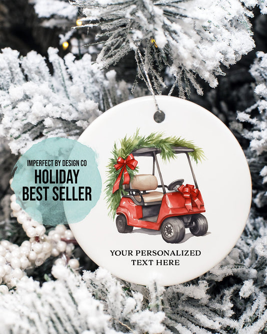 imperfect by design co design exclusive 
A round white Christmas ornament featuring a red golf cart decorated with green pine garlands and a red bow, hanging on a frosted Christmas tree. A teal circular badge on the left reads 'IMPERFECT BY DESIGN CO HOLIDAY BEST SELLER' and the ornament includes placeholder text 'YOUR PERSONALIZED TEXT HERE'.