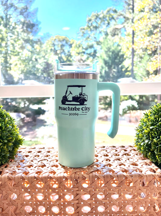 Exclusive imperfect by design co design on 20oz Turquoise Mini Tumbler reads 'Peachtree City' 'Est. 1959' '30269' placed on rattan/wicker surface flanked by to boxwood proprieties with trees visible through window in background