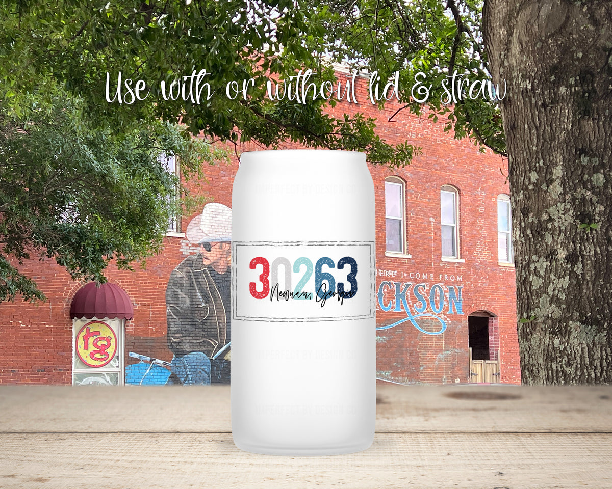 A frosted  tumbler with a printed '30263 Newnan, Georgia' design in red, white, and blue, shown on a wooden surface with a brick building and trees in the background. White script text at the top reads 'Use with or without lid & straw'.