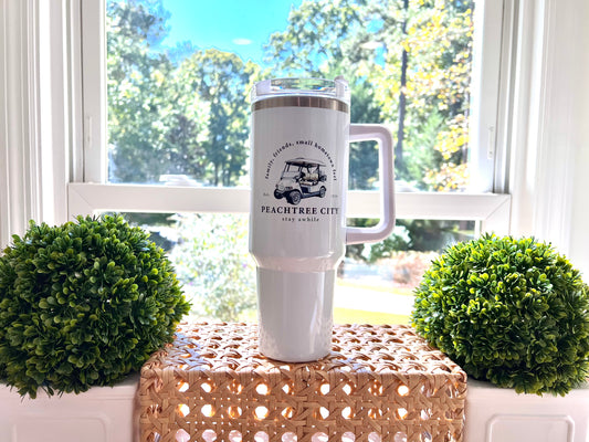 imperfect by design co exclusive White tumbler with white golf cart and text placed on woven surface in front of a window with a view of trees, flanked by two boxwood topiaries.