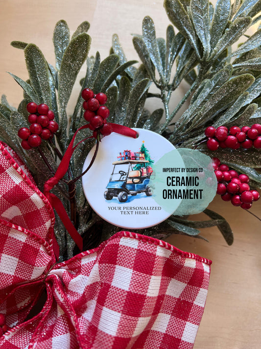 A close-up of a round ceramic holiday ornament attached to a red ribbon, displayed among green frosted pine branches, red berry clusters, and a red-and-white gingham bow. The ornament features an illustration of a golf cart carrying Christmas presents, with text reading ''YOUR PERSONALIZED TEXT HERE"