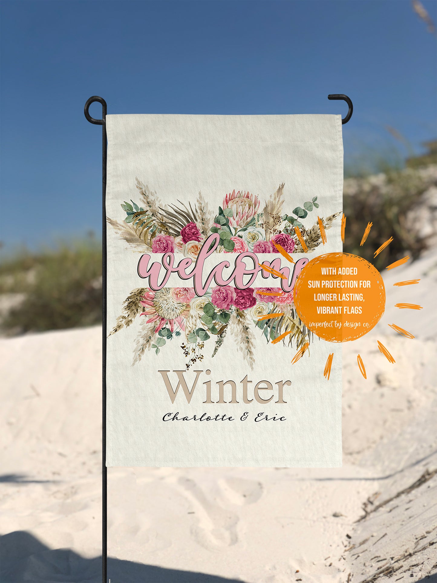 Decorative garden flag with floral design and text 'Welcome' & Personalized with 'Winter, Charlotte & Eric' on a beach background. Sun shaped text graphic reads "with added protection for longer lasting vibrant flags imperfect by design co'