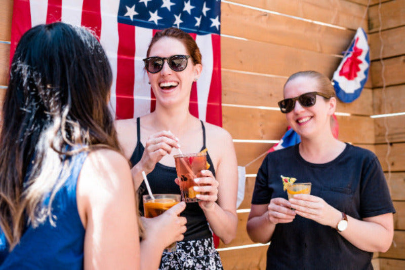 Three women laughing and holding cups of sweet southern ice tea in front of a rustic wooden wall with an American flag hanging behind them. The central woman wears a black tank top and sunglasses, while the woman on the right wears a black t-shirt and sunglasses.