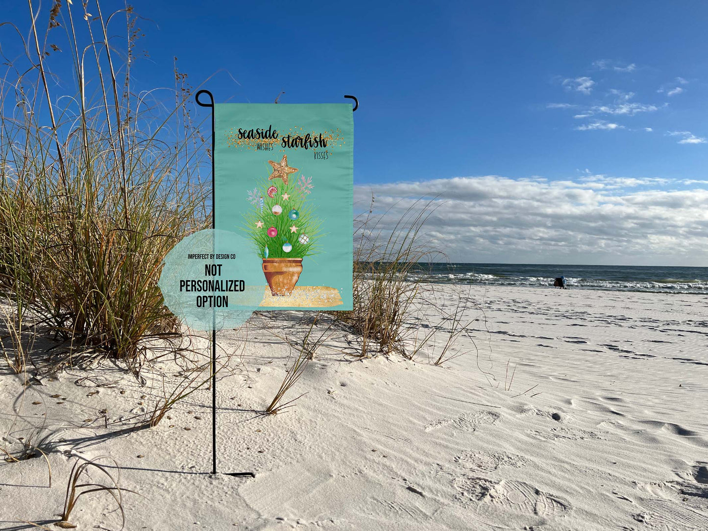 Decorative not personalized flag option. The blue green colored garden flag featuring potted ornamental grass adorned with Christmas tree bulb and topped with a golden starfish. The Flag reads 'Seaside Wishes Starfish Kisses'. Large golden brushstroke below the plant. The flag is on a flag pole. The garden flag is hung on a garden flag pole with beach  on a sandy beach with ocean view. Large blue blurb is placed on the graphic that reads 'imperfect by design co'  'not personalized option'.
