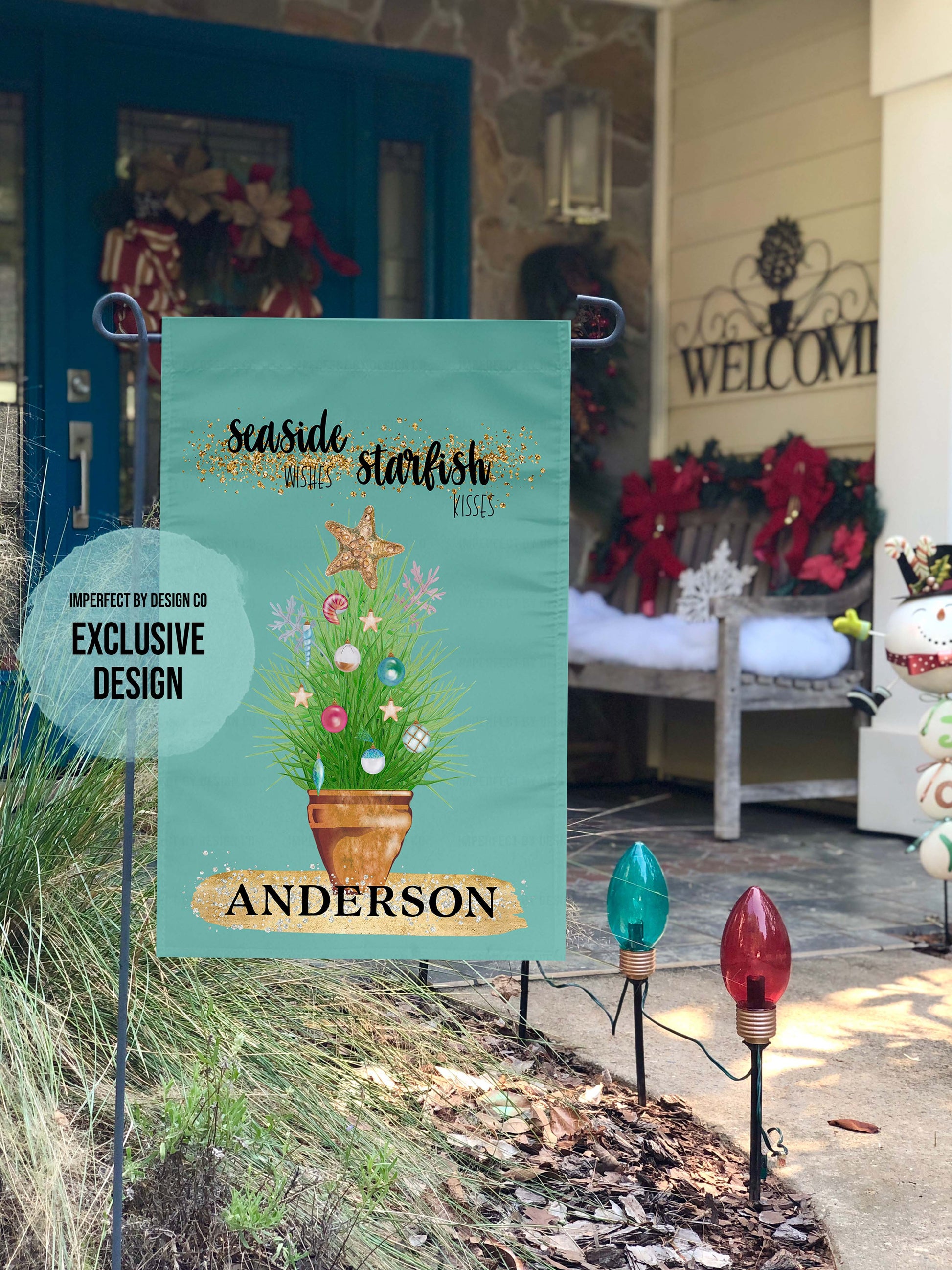Personalized with a Last Name Flag. The blue green colored garden flag featuring potted ornamental grass adorned with Christmas tree bulb and topped with a golden starfish. The Flag reads 'Seaside Wishes Starfish Kisses'. Below the potted plant there is a large golden brushstroke with black bold text that reads 'Anderson'. The flag is on a flag pole. A holiday decorate porch is visible in the background.  A blue blurb on the flag reads imperfect by design co exclusive design.