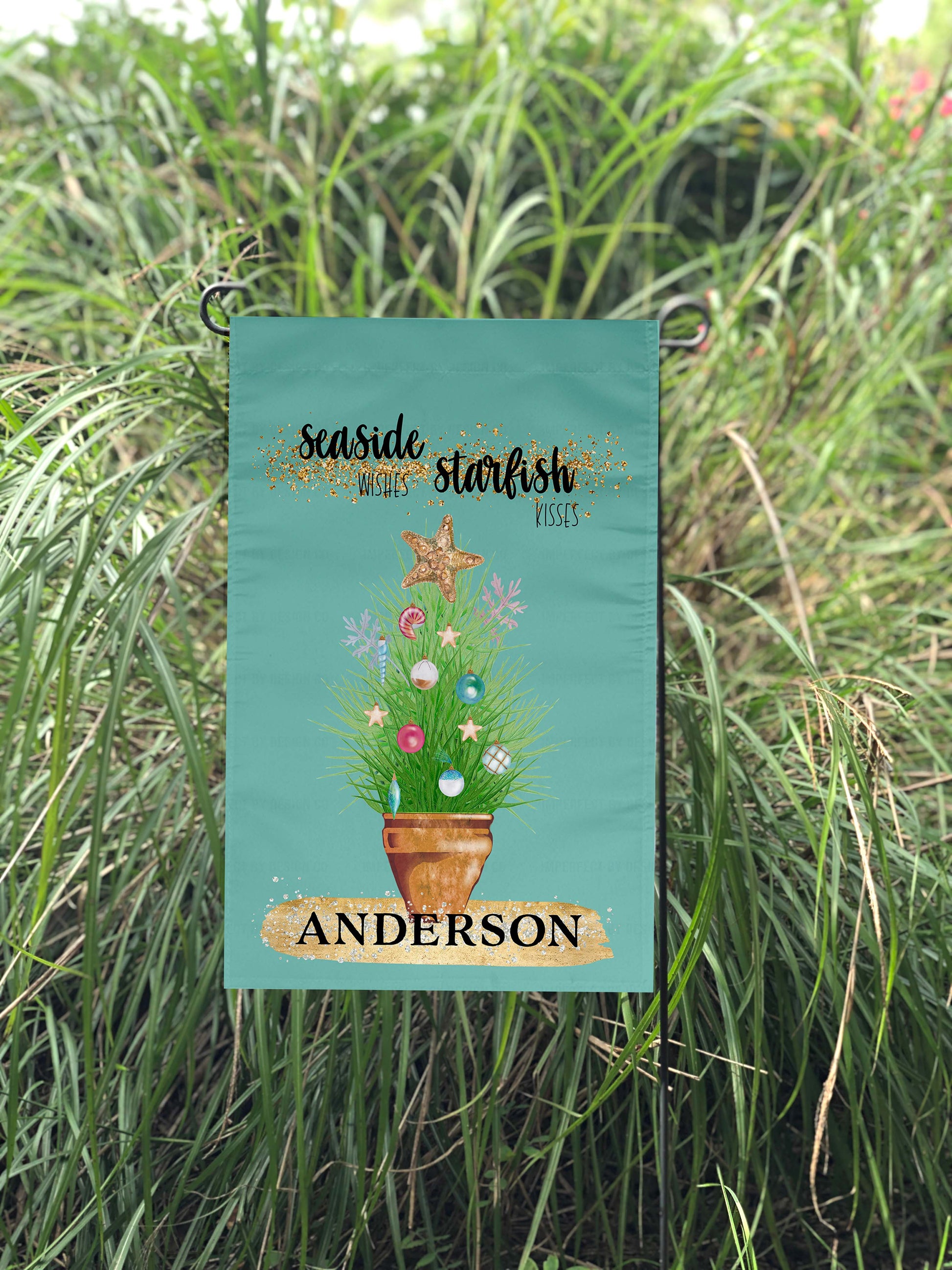 Personalized with a Last Name Flag. The blue green colored garden flag featuring potted ornamental grass adorned with Christmas tree bulb and topped with a golden starfish. The Flag reads 'Seaside Wishes Starfish Kisses'. Large golden brushstroke below the plant with black bold text that reads 'Anderson. The flag is on a flag pole. Large Ornamental Grass is visible in the background.