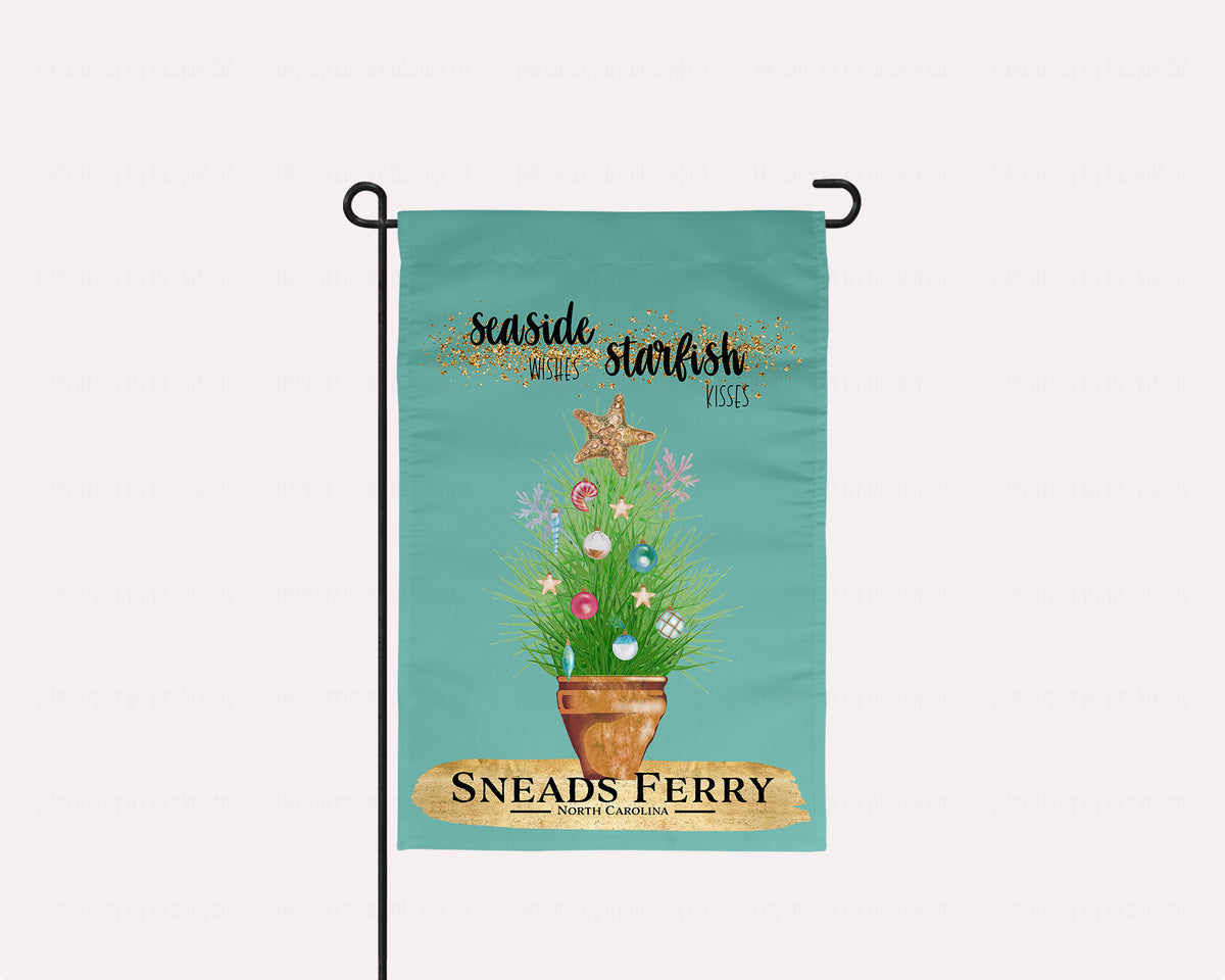 imperfect by design co exclusive Coastal Vibes Collection. Flag features City & State Personalization. The blue green colored garden flag featuring potted ornamental grass adorned with Christmas tree bulb and topped with a golden starfish. The Flag reads 'Seaside Wishes Starfish Kisses'. Below the potted plant there is a large golden brushstroke with black bold text that reads 'Sneads Ferry' 'North Carolina' The flag is on a flagstand placed on a pale pink background.