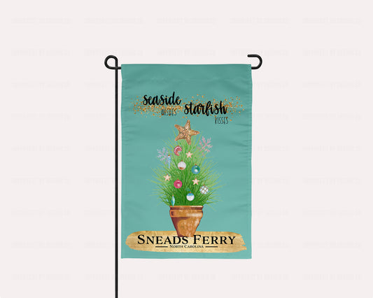 imperfect by design co exclusive Coastal Vibes Collection. Flag features City & State Personalization. The blue green colored garden flag featuring potted ornamental grass adorned with Christmas tree bulb and topped with a golden starfish. The Flag reads 'Seaside Wishes Starfish Kisses'. Below the potted plant there is a large golden brushstroke with black bold text that reads 'Sneads Ferry' 'North Carolina' The flag is on a flagstand placed on a pale pink background.