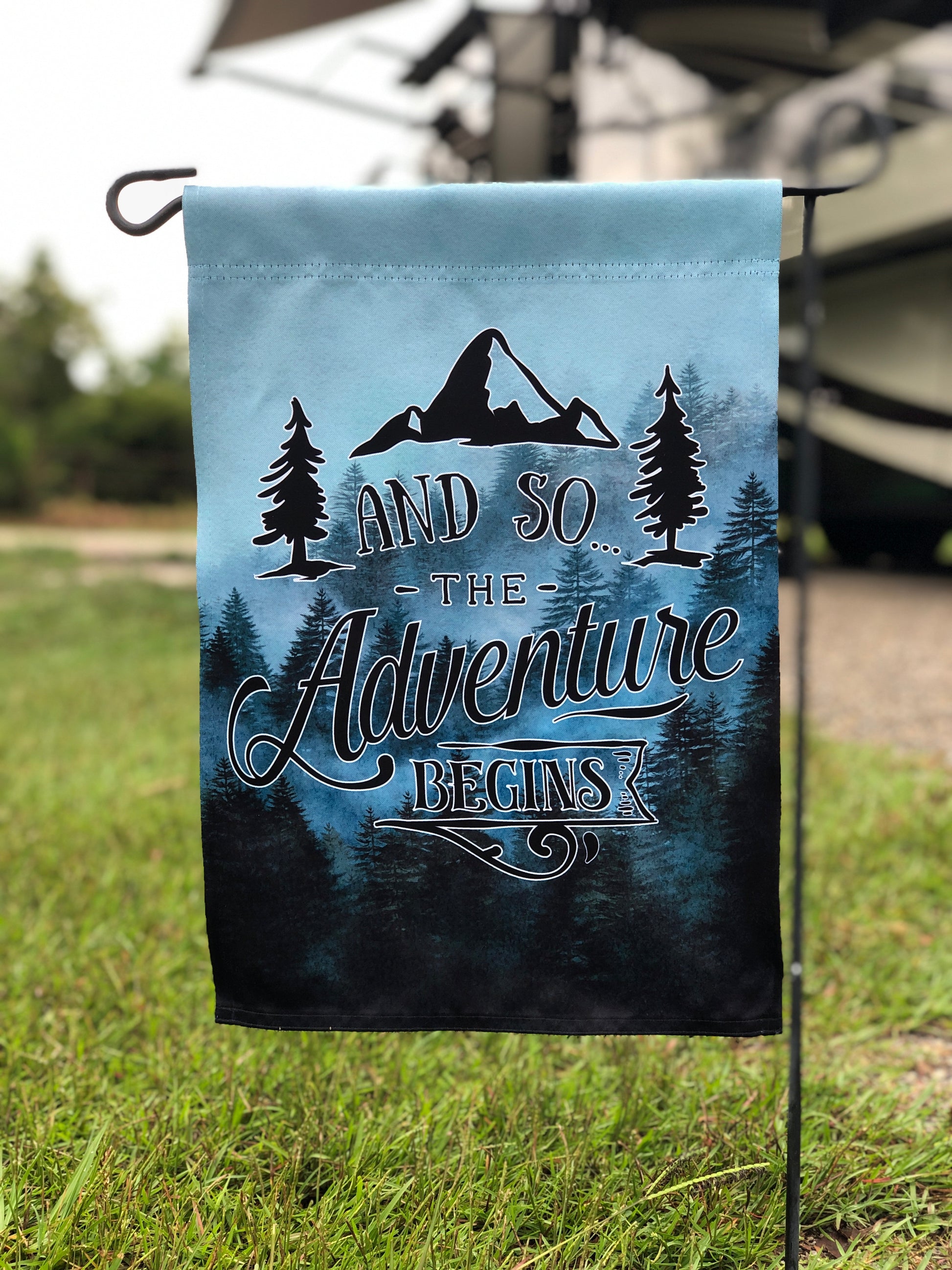 No Personalized Option 12x20 inch Garden flag with moody mountain and tree scene, text reads 'And so the adventure begins' Flag is placed on a flag stand on a grassy patch in front of an RV