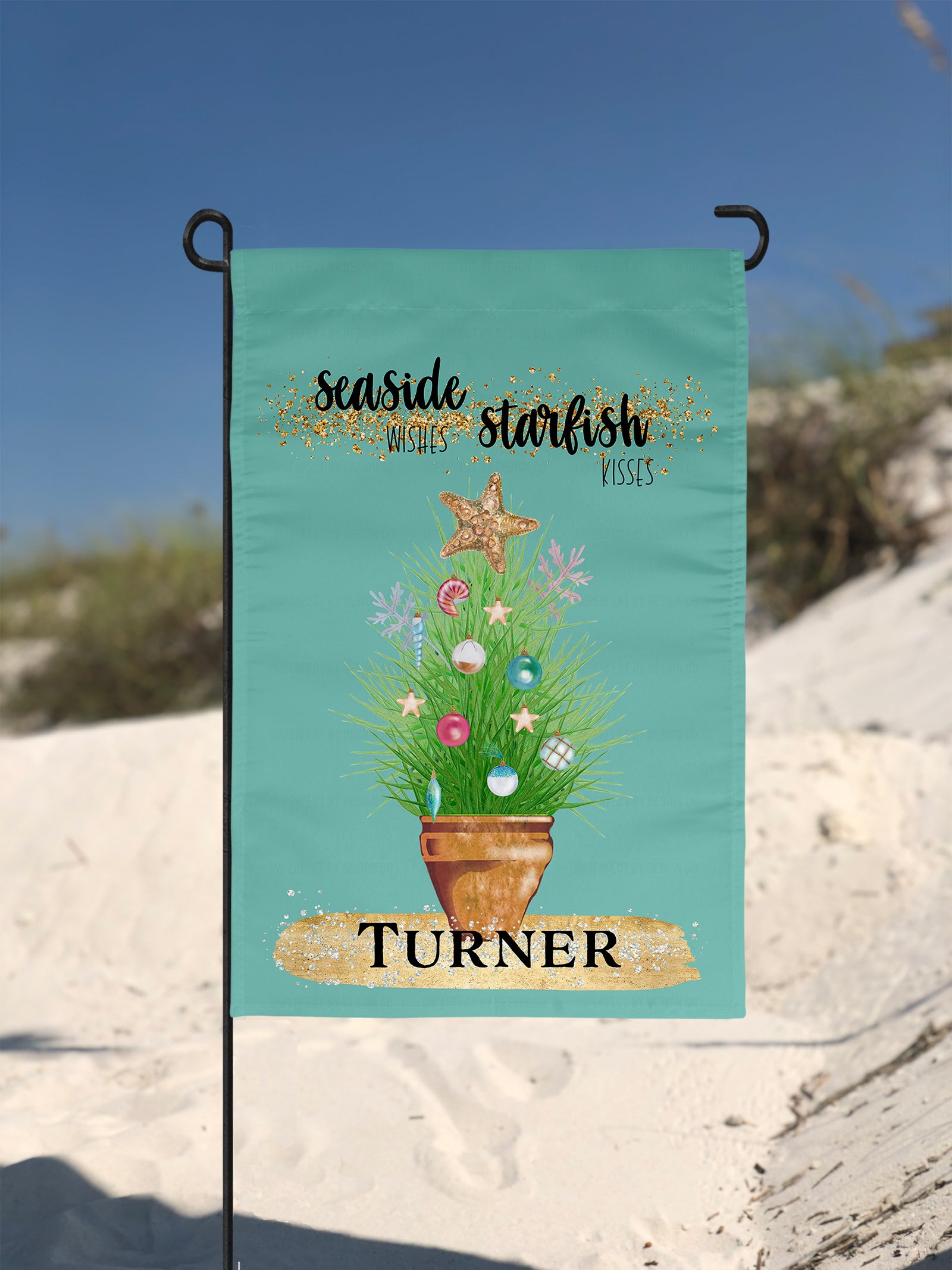 Personalized with a Last Name Flag. The blue green colored garden flag featuring potted ornamental grass adorned with Christmas tree bulb and topped with a golden starfish. The Flag reads 'Seaside Wishes Starfish Kisses'. Below the potted plant there is a large golden brushstroke with black bold text that reads 'Turner'. The flag is on a flag pole. A sand dune is visible in the background,
