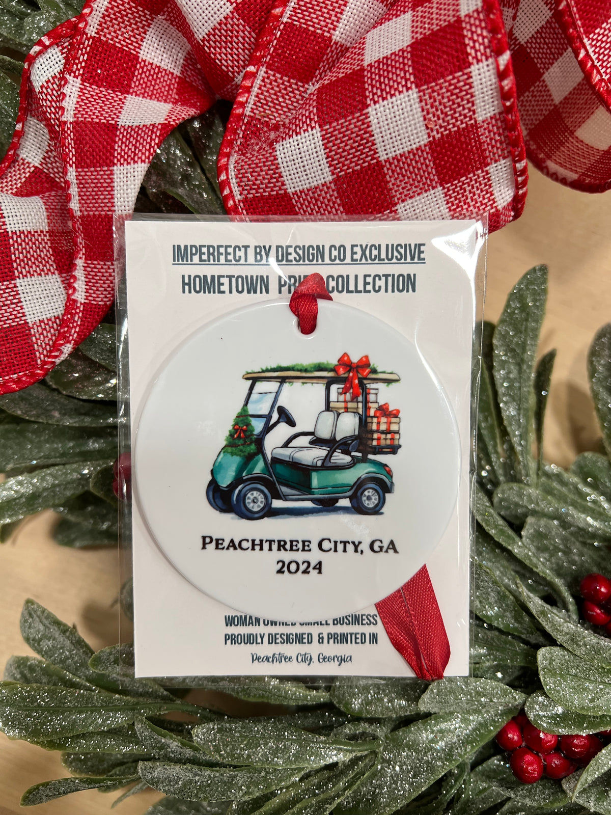 imperfect by design co exclusive design 
Glossy white ornament with a green golf cart, personalized with custom city and state or family name, complete with a red ribbon. Text on ornament reads "Peachtree City 2024". The ornament is package in clear branded cellophane placed on top of a green wreath with red berries and a red bow.