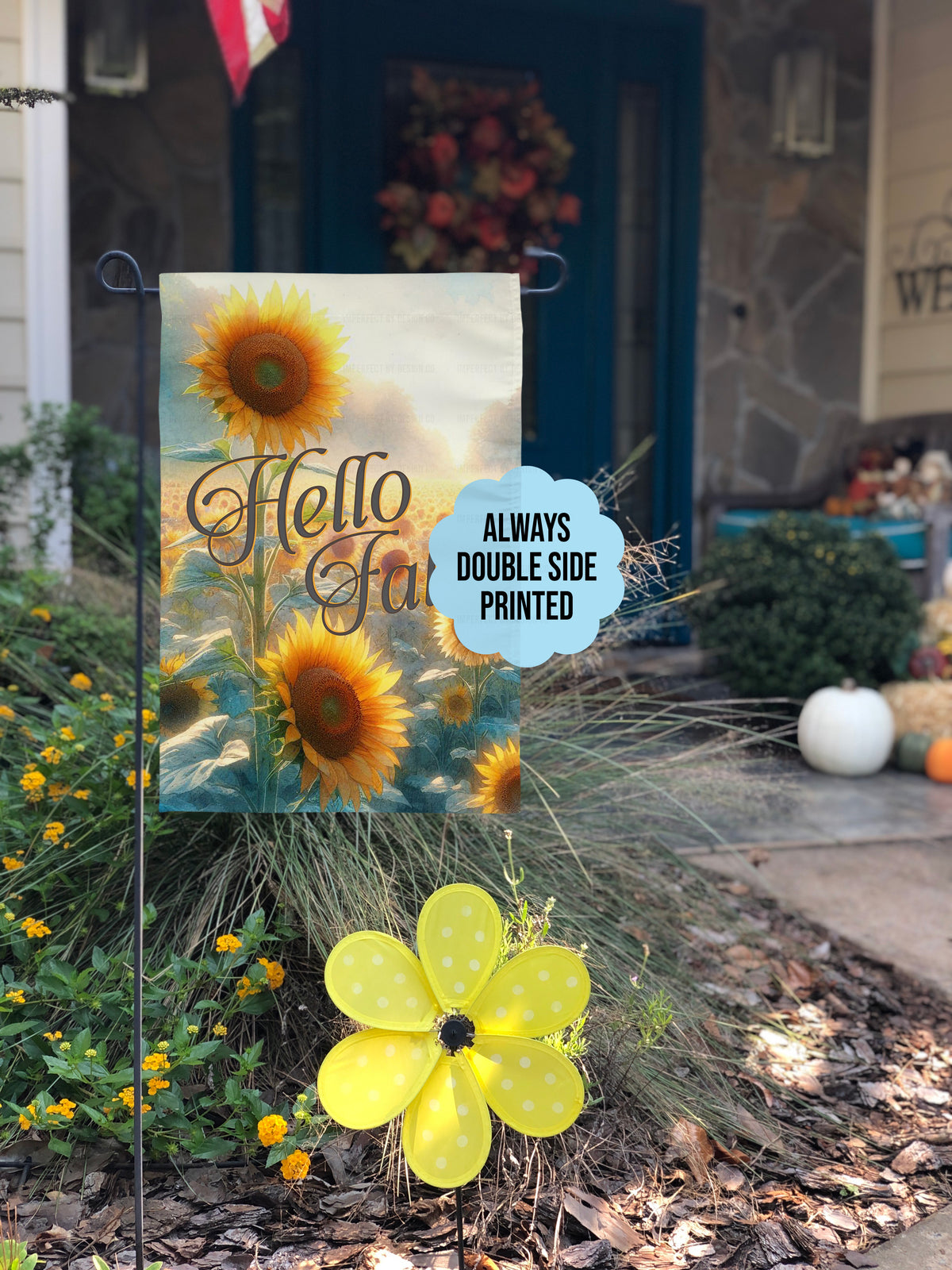 A decorative garden flag with sunflower artwork and the words 'Hello Fall' is displayed outdoors in a front yard beside a yellow flower wind spinner. A light blue cloud-shaped sticker reads 'ALWAYS DOUBLE SIDE PRINTED'.