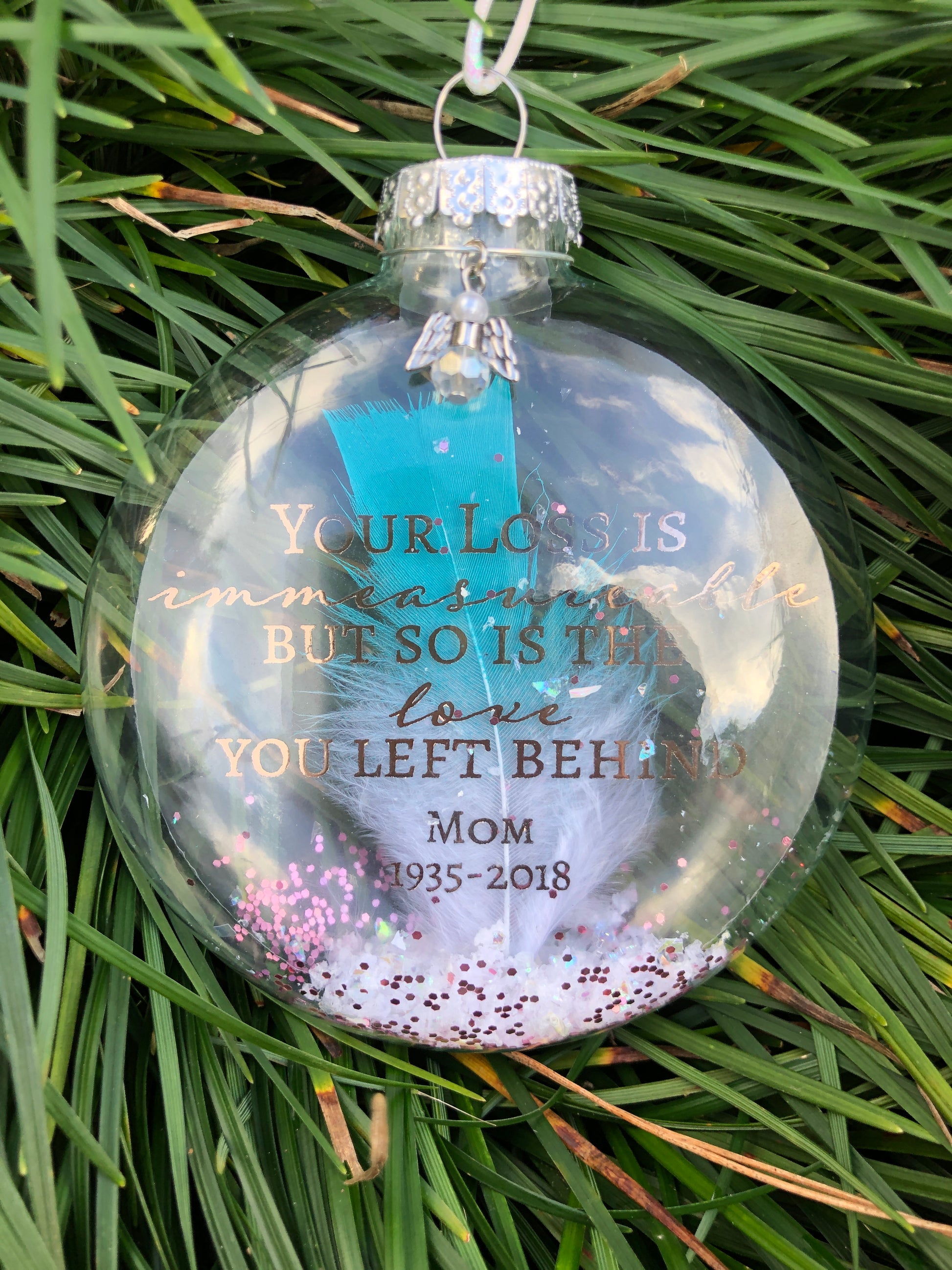 Glass memorial ornament featuring a white feather with teal tip and angel charm.