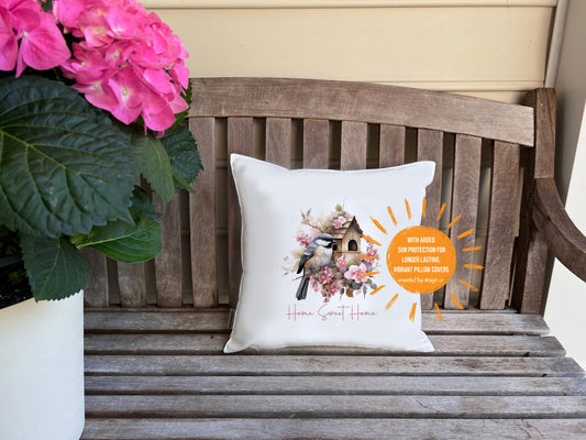 Decorative pillow (cover) with floral and bird design on a wooden bench flanked by pink hydrangea flowers. Sun shaped text blurb reads 'with added sun protection for longer lasting vibrant pillow covers imperfect by design co'
