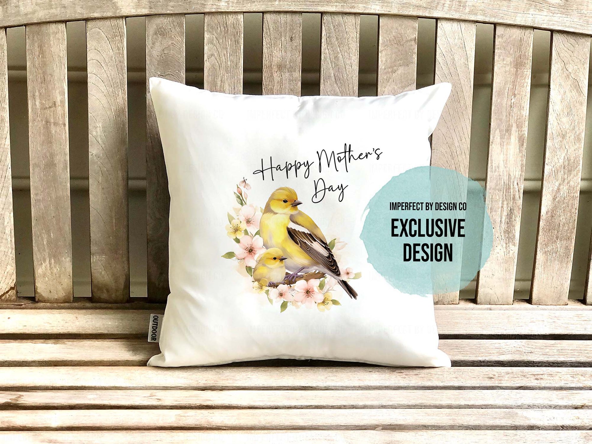 White dcorative white pillow advertising pillow cover with a female Goldfinch and its nestling on floral branch. The pillow is placed on a teak bench. The text on the pillow reads 'Happy Mother's Day'. Teal text blurb on pillow reads 'imperfect by design co exclusive design'
