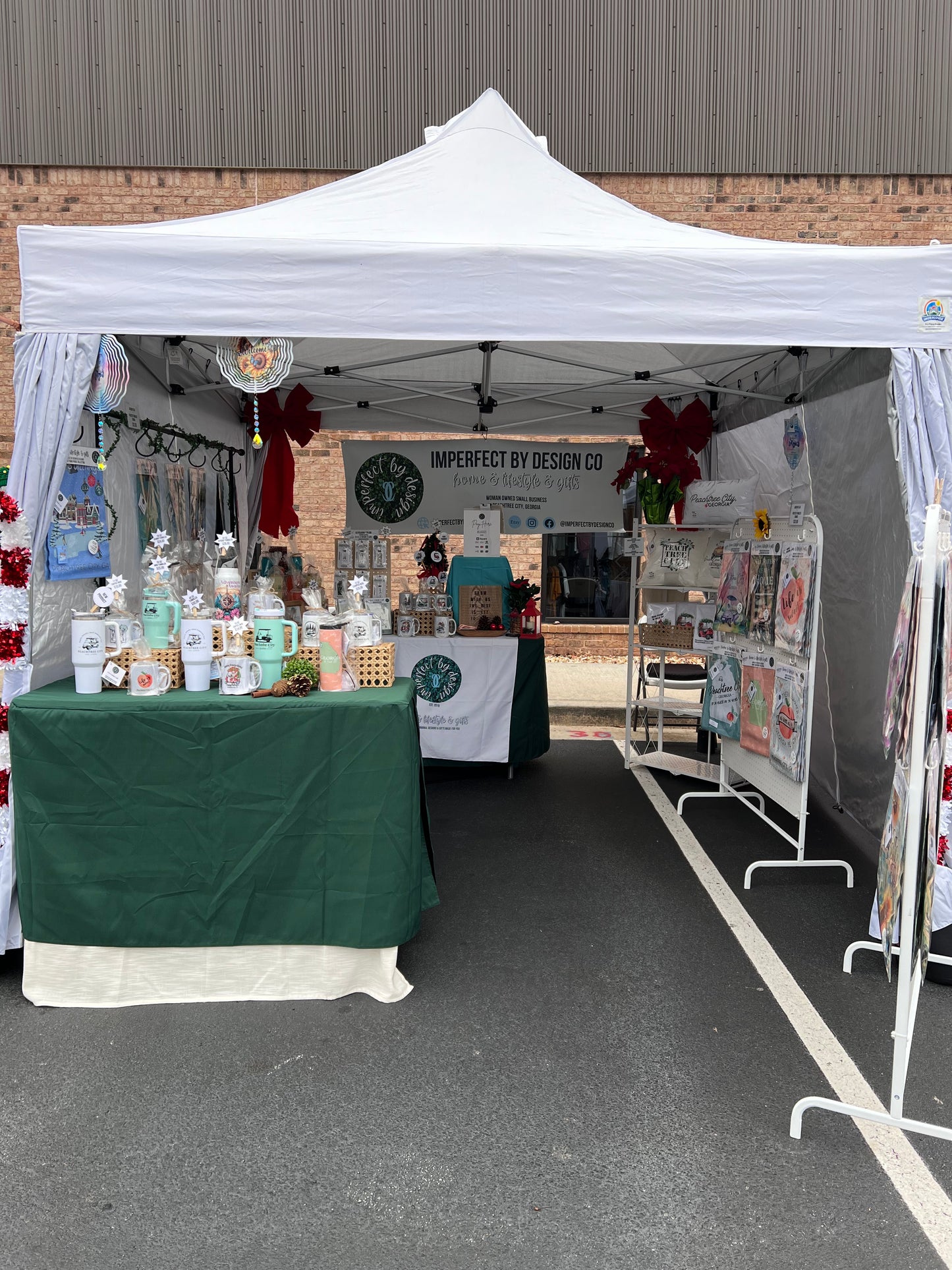 White vendor tent and tables with green table cloth. Items on tables range from drinkware to keychains and holiday ornaments. Large banner in background reads imperfect by design co home & lifestyle & gifts as well as a large imperfect by design co logo. Colorful garden flags, pillows and wind spinners are also visible. The vendor text is decorated for the winter holidays